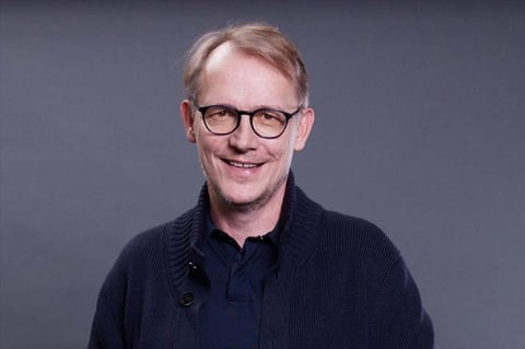 Middle-aged man with reddish-brown hair and glasses smiling at camera against gray background